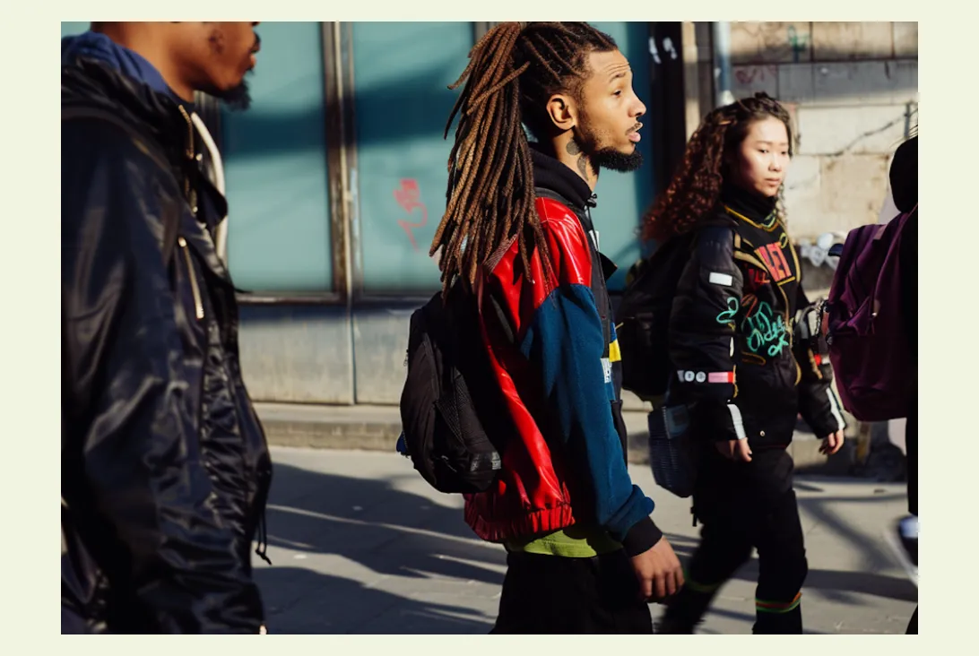 Three young adults walking outdoors in sunlight wearing backpacks and colorful jackets, one with long dreadlocks.