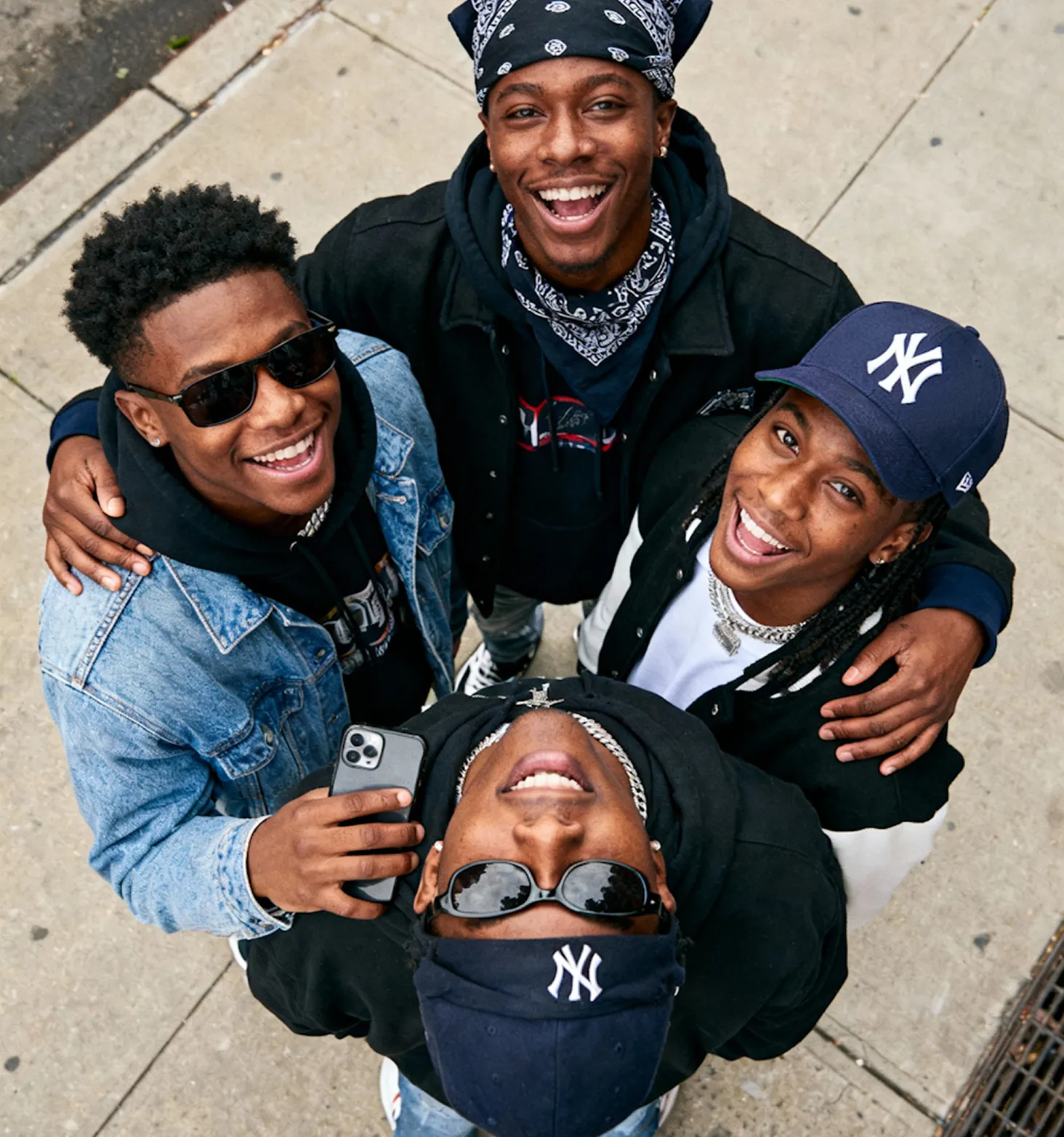 Four young men standing close together on a sidewalk, smiling and looking up at the camera, two wearing New York Yankees caps.