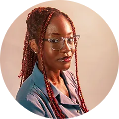 Portrait of a woman with long red braided hair, glasses, and a blue shirt against a beige background.