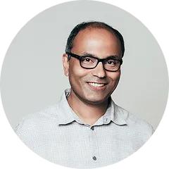 Smiling man with glasses wearing a light gray button-up shirt against a plain light background.