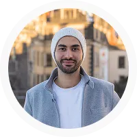 Man with beard wearing white beanie and gray jacket standing outdoors in blurred urban background.
