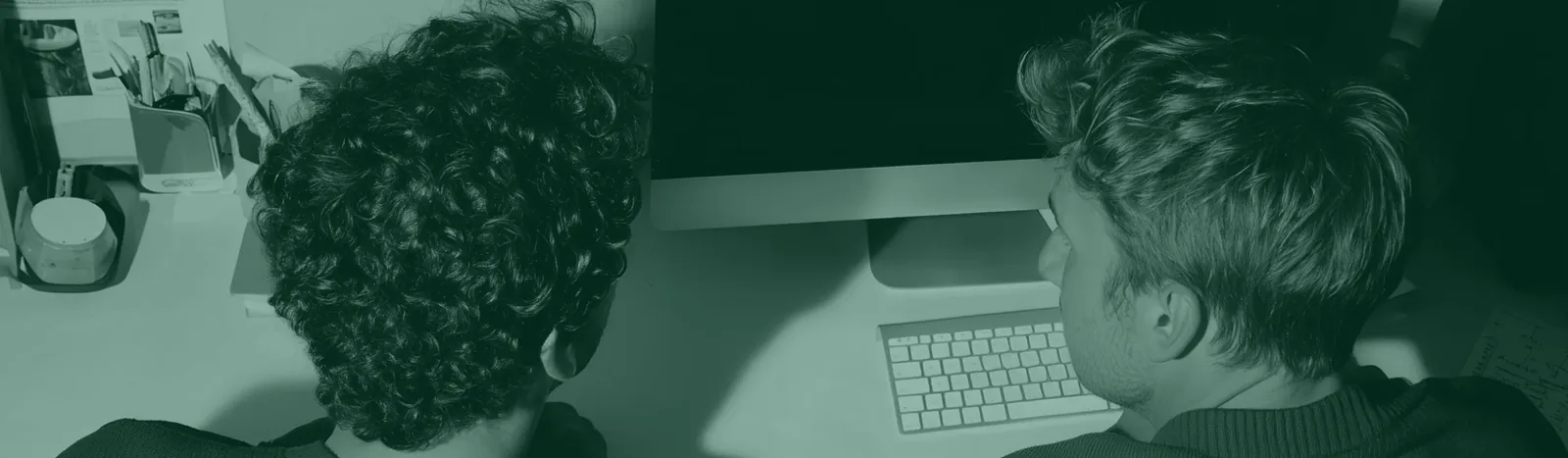 Two people with curly and straight hair working together at a desk with a computer and keyboard.